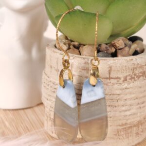 Boucles d’oreilles pendantes en opale bleue naturelle avec crochets dorés hypoallergéniques, posées devant une plante.